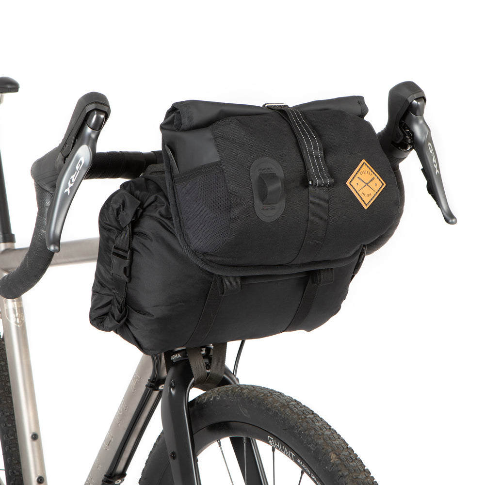 A black Restrap Holster Bar Bag Plus mounted on a bicycle handlebar, featuring a rolltop canister bag attached in front.