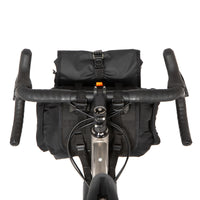 A black Restrap Holster Bar Bag Plus mounted on a bicycle handlebar, featuring a rolltop canister bag attached in front, shown from behind and above