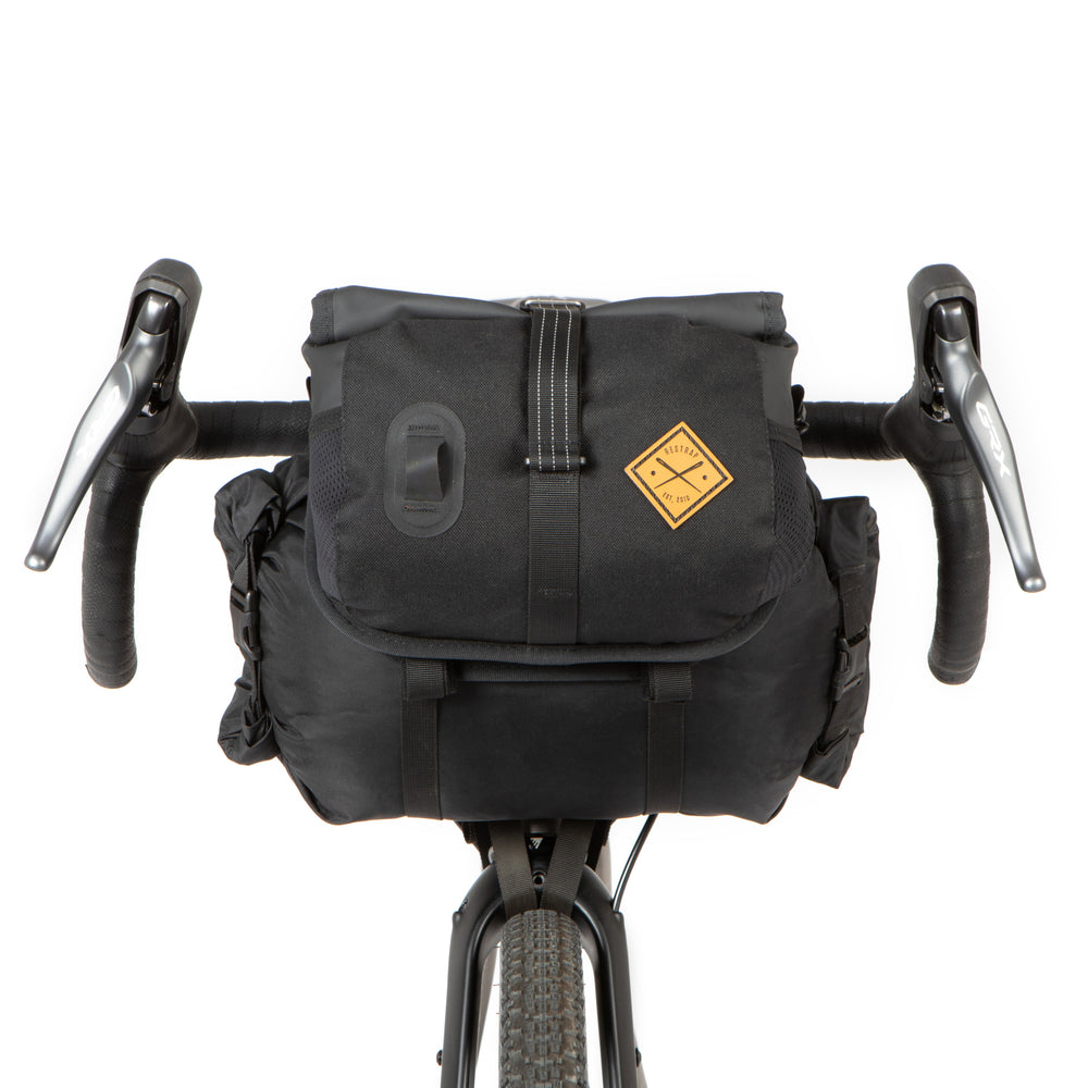 A black Restrap Holster Bar Bag Plus mounted on a bicycle handlebar, featuring a rolltop canister bag attached in front.