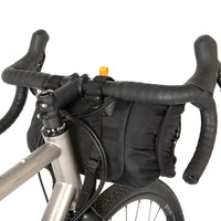 A black restrap holster bar bag attached to the handlebars of a bicycle, designed for bikepacking, shown from the rear