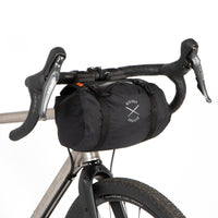 A black Race Bar Bag attached to the handlebars of a bicycle, featuring a holster system and a roll-top dry bag - this is attached to drop handlebars