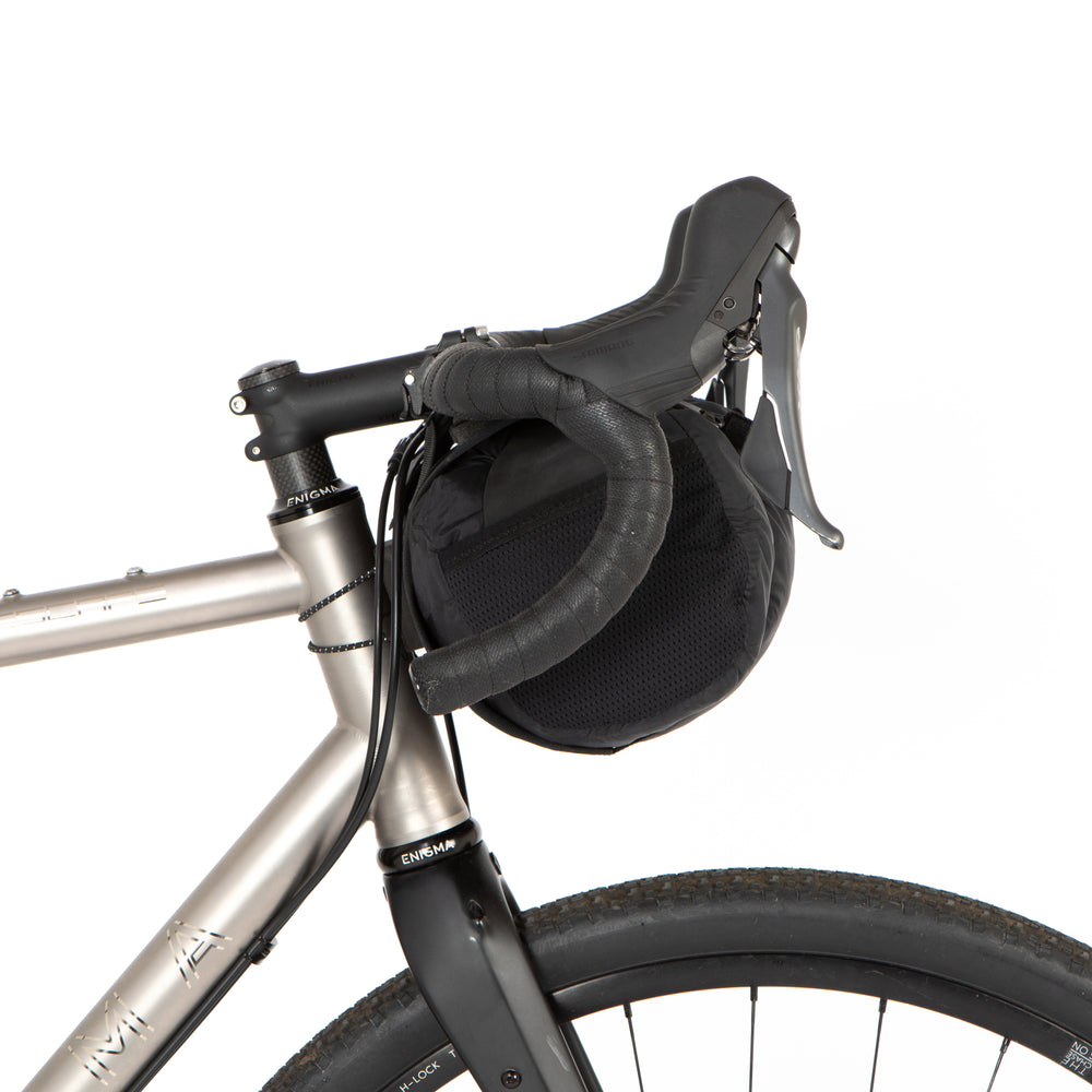 A black Race Bar Bag attached to the handlebars of a bicycle, featuring a holster system and a roll-top dry bag - this is attached to drop handlebars