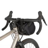 A black Race Bar Bag attached to the handlebars of a bicycle, featuring a holster system and a roll-top dry bag - this is attached to drop handlebars