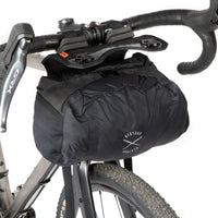A black Race Bar Bag attached to the handlebars of a bicycle, featuring a holster system and a roll-top dry bag - this is attached to drop handlebars