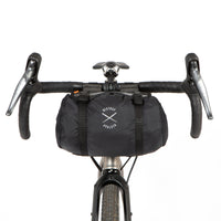 A black Race Bar Bag attached to the handlebars of a bicycle, featuring a holster system and a roll-top dry bag - this is attached to drop handlebars