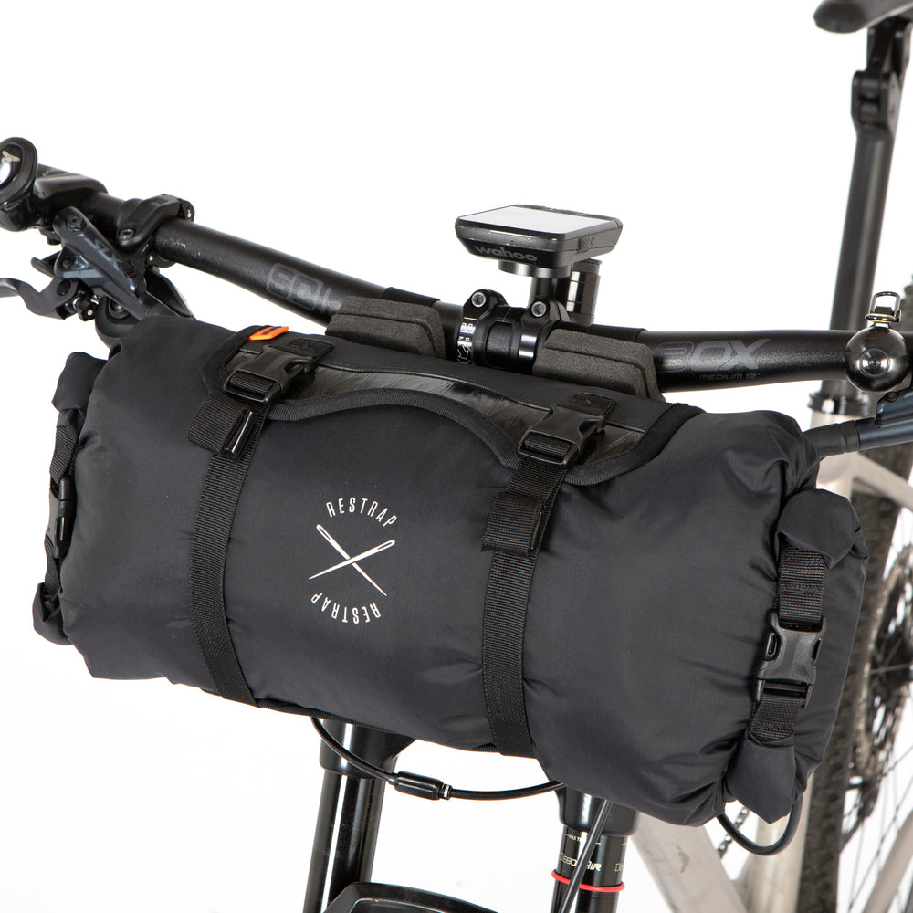 A black Race Bar Bag attached to the handlebars of a bicycle, featuring a holster system and a roll-top dry bag - this is attached to flat handlebars on a mountain bike