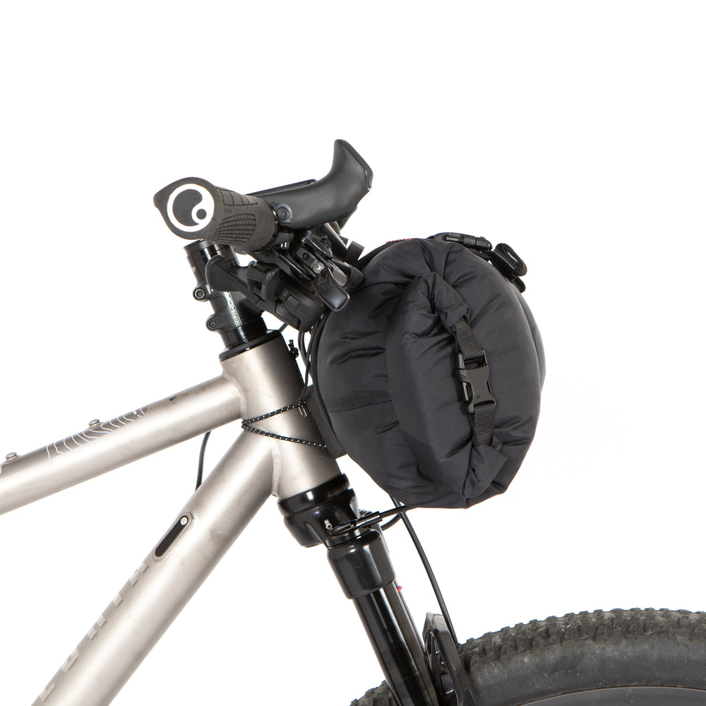A black Race Bar Bag attached to the handlebars of a bicycle, featuring a holster system and a roll-top dry bag - this is attached to flat handlebars on a mountain bike