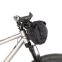 A black Race Bar Bag attached to the handlebars of a bicycle, featuring a holster system and a roll-top dry bag - this is attached to flat handlebars on a mountain bike