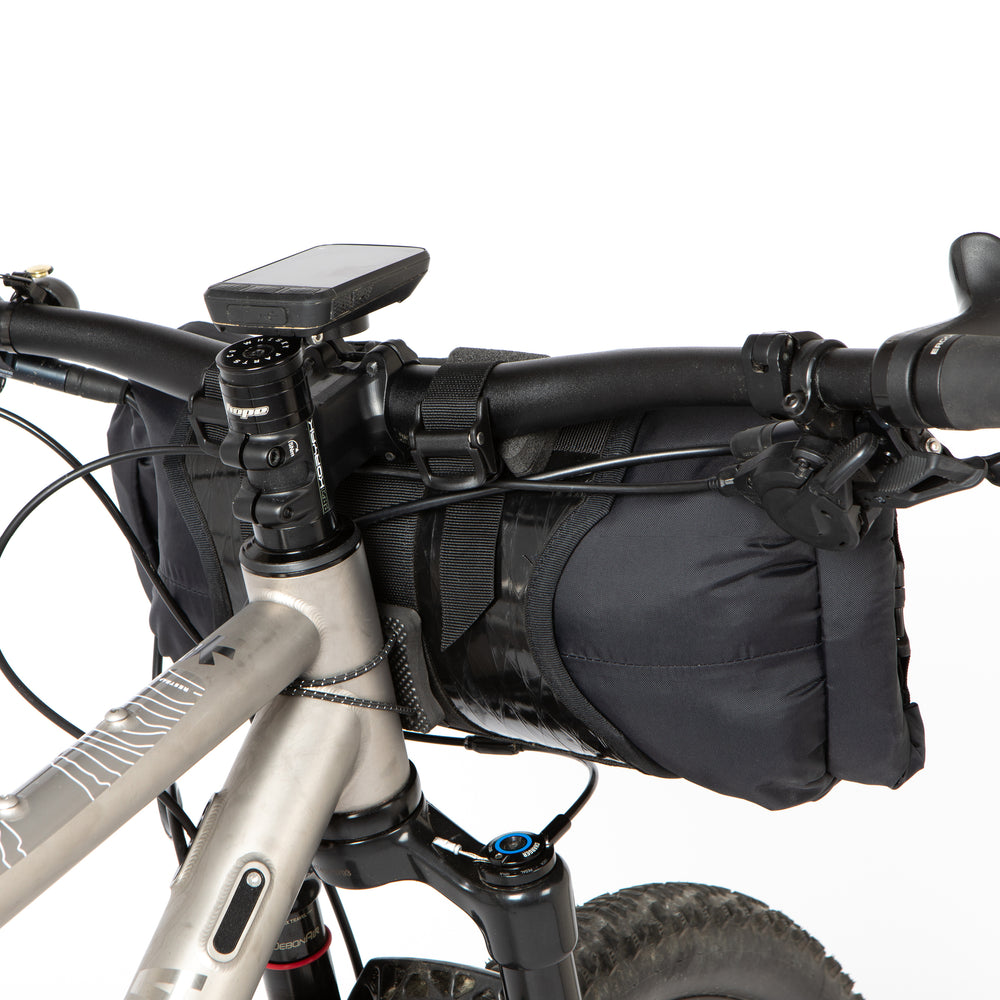 A black Race Bar Bag attached to the handlebars of a bicycle, featuring a holster system and a roll-top dry bag - this is attached to flat handlebars on a mountain bike