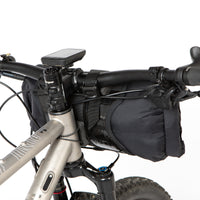 A black Race Bar Bag attached to the handlebars of a bicycle, featuring a holster system and a roll-top dry bag - this is attached to flat handlebars on a mountain bike