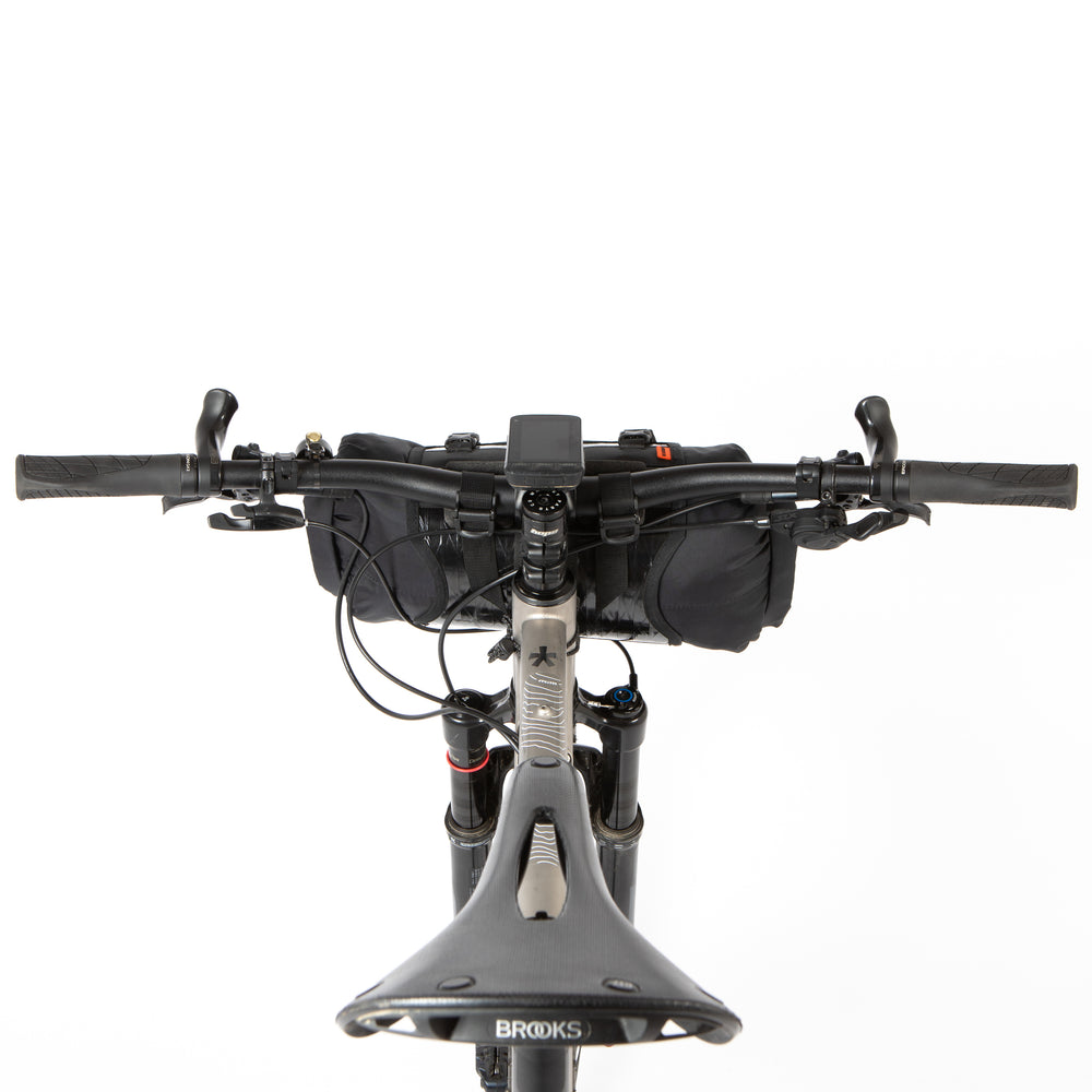 A black Race Bar Bag attached to the handlebars of a bicycle, featuring a holster system and a roll-top dry bag - this is attached to flat handlebars on a mountain bike