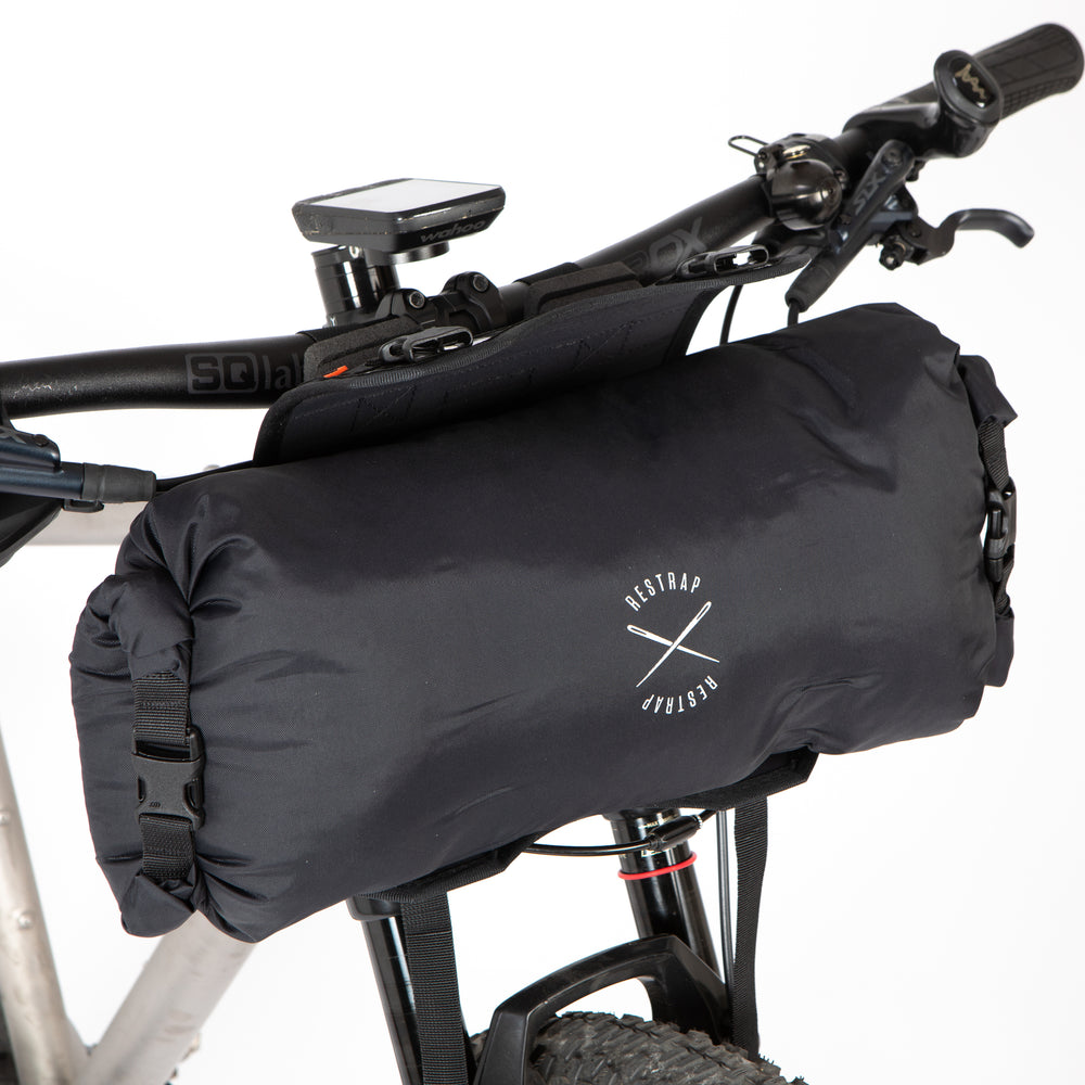 A black Race Bar Bag attached to the handlebars of a bicycle, featuring a holster system and a roll-top dry bag - this is attached to flat handlebars on a mountain bike. The dry bag is being removed from the holster