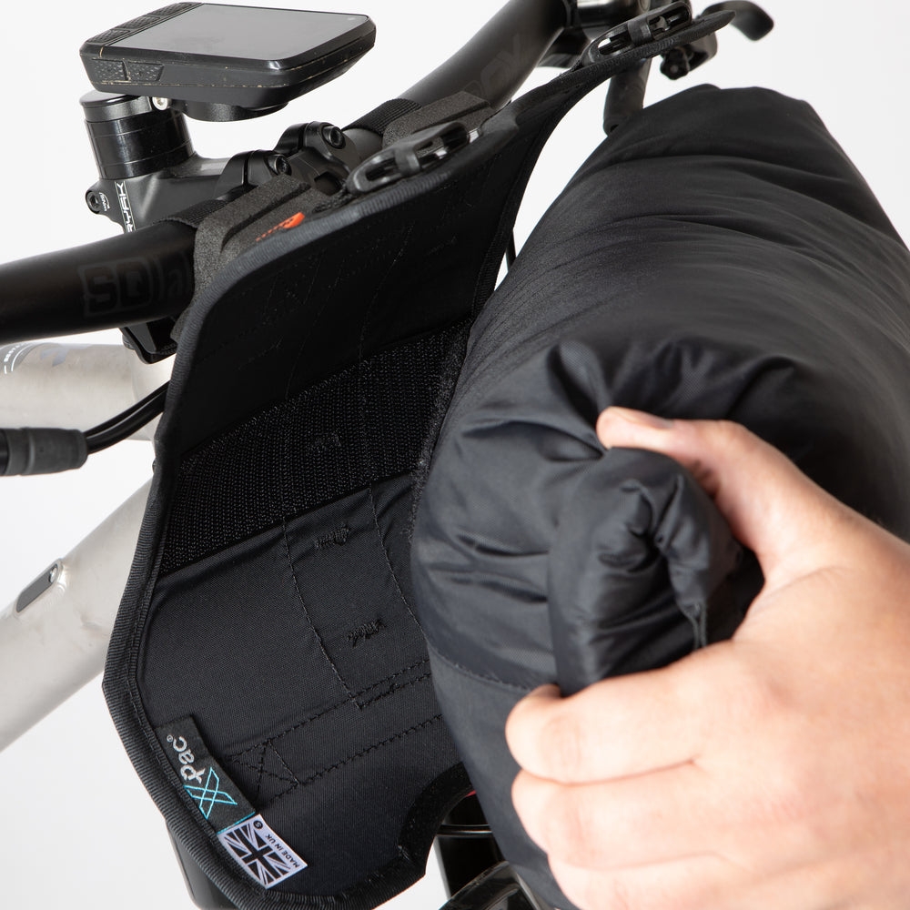 A black Race Bar Bag attached to the handlebars of a bicycle, featuring a holster system and a roll-top dry bag - this is attached to flat handlebars on a mountain bike. The dry bag is being removed from the holster