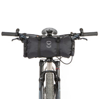 A black Race Bar Bag attached to the handlebars of a bicycle, featuring a holster system and a roll-top dry bag - this is attached to flat handlebars on a mountain bike