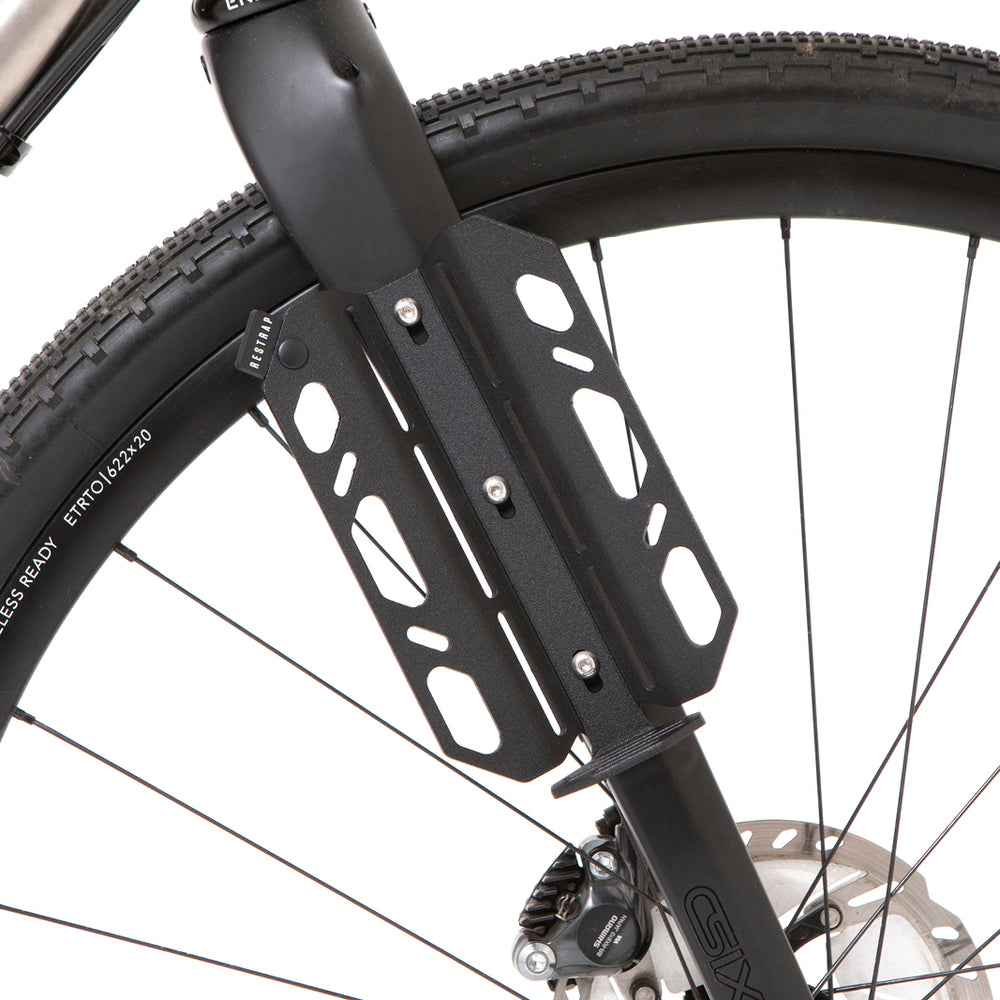 A black laser cut aluminium cargo cage mounted to the fork of a bicycle, with Restrap branded rubber label visible.