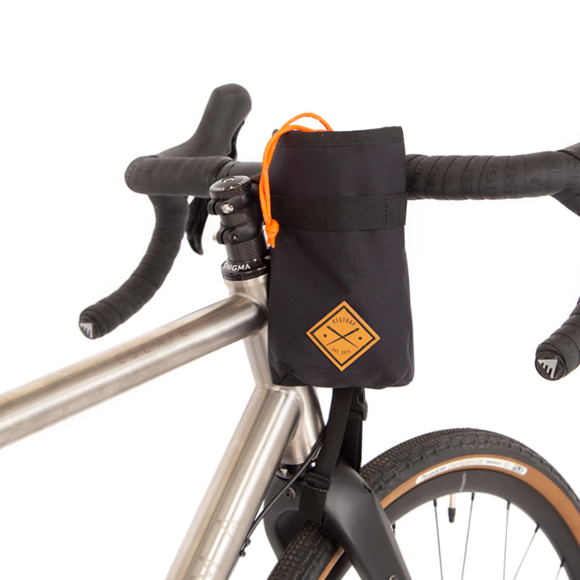 A black Restrap stem bag mounted on the handlebar of a bicycle, designed for carrying a water bottle or other items, with an orange drawstring and the Restrap logo in yellow.