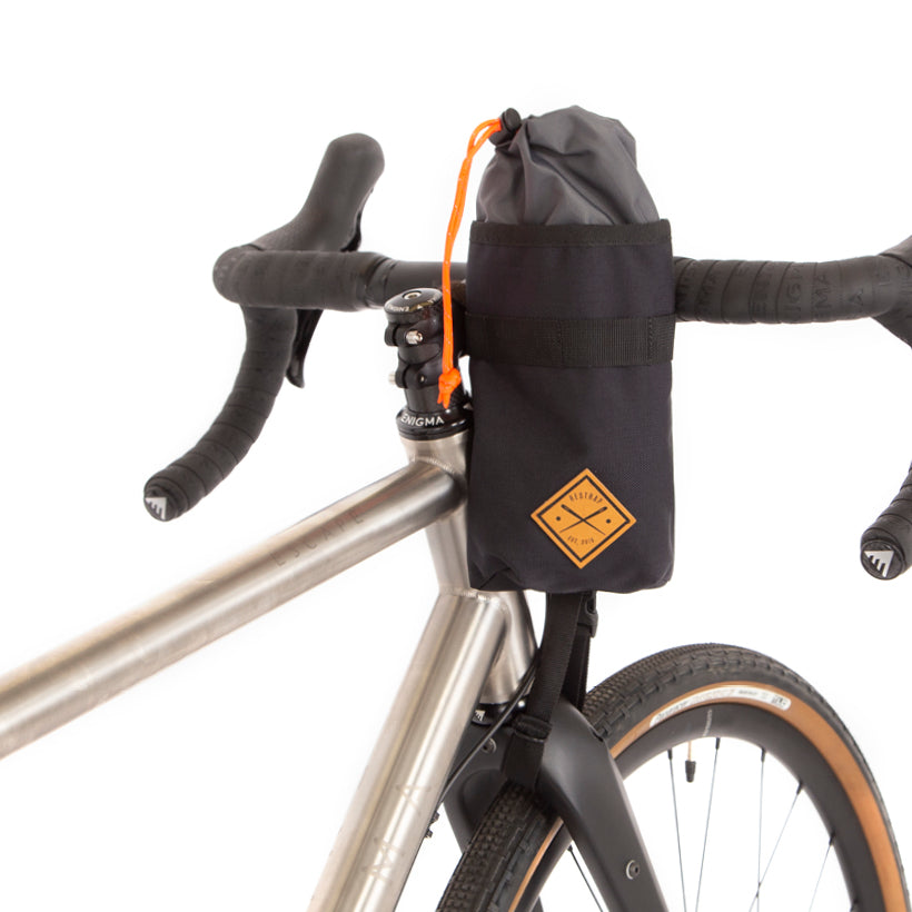 A black Restrap stem bag mounted on the handlebar of a bicycle, designed for carrying a water bottle or other items, with an orange drawstring and the Restrap logo in yellow.