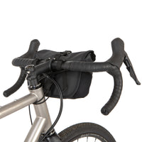 A black Rolltop Canister Bag attached to the handlebars of a bicycle, shown from the rear